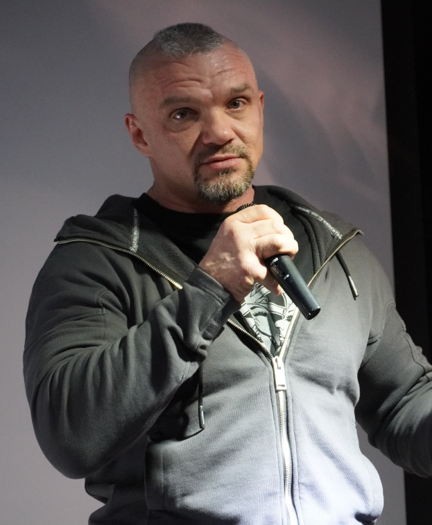 Ein Mann in einer schwarzen Kapuzenjacke und einem schwarzen Anzug mit einem wei├čen Hemd, der ein Mikrofon h├Ąlt, steht selbstbewusst vor einer Wand und scheint ein Komiker zu sein.