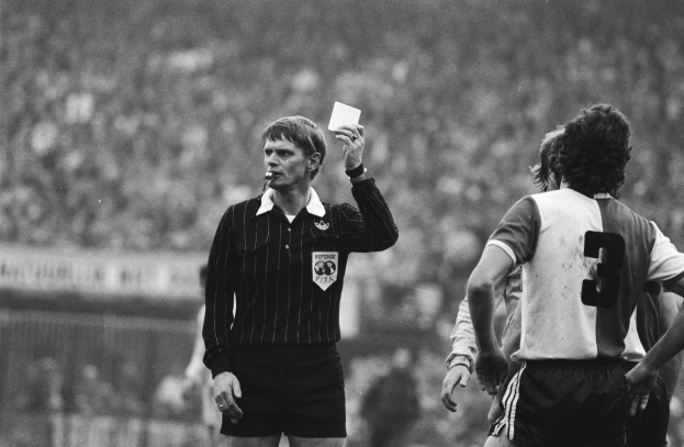 Ein Schiedsrichter in Schwarz-Weiß-Bild hält eine Rote Karte vor einer Menge von Menschen in Fußballtrikots, mit einem Zaun im Hintergrund.