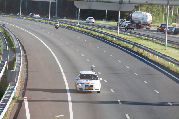 Ein Auto auf einer Autobahn mit einem stationären Polizeiwagen am Rande, andere Fahrzeuge in Bewegung und Straßeninfrastruktur einschließlich Geländern, Pfosten, Schildern und Bäumen.