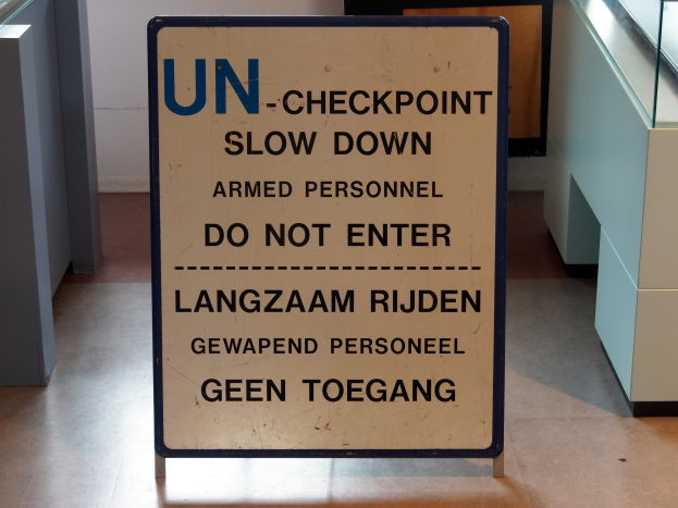 Schild auf dem Boden mit der Aufschrift "UN-Checkpoint Verlangsame bewaffnete Person - Betreten verboten" mit einem Glastisch und Gegenständen rechts daneben und einer Wand mit einem Gegenstand im Hintergrund.