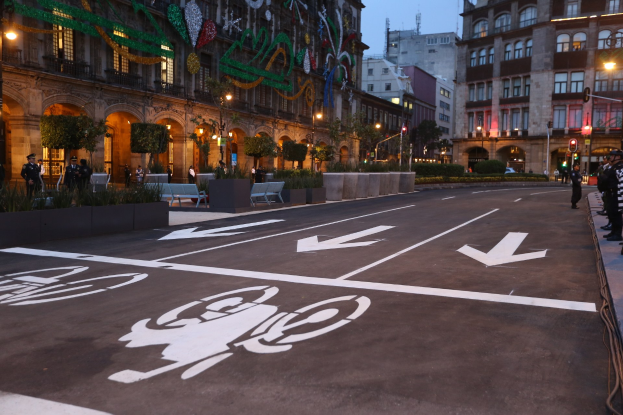 Stadtstraße mit einem Fahrradweg, umgeben von Gebäuden, Straßenmöbeln, Fußgängern, Grünflächen und einem klaren Himmel.