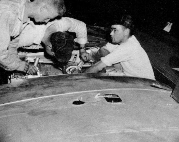 Drei Männer mit Mützen arbeiten an einem schwarzen und weißen Auto in einer Garage, umgeben von Werkzeugen und Materialien.