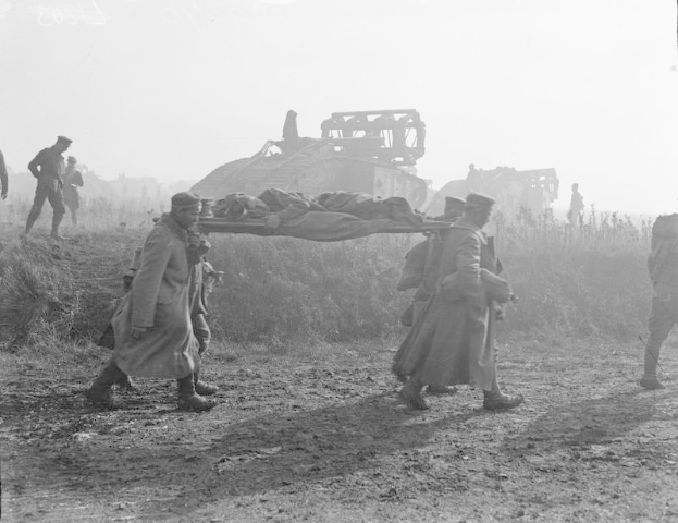 Schwarzes und weißes Bild von Männern, die einen verletzten Mann auf einer Trage eine Schotterstraße entlang tragen, mit einem Panzer und Vegetation im Hintergrund.