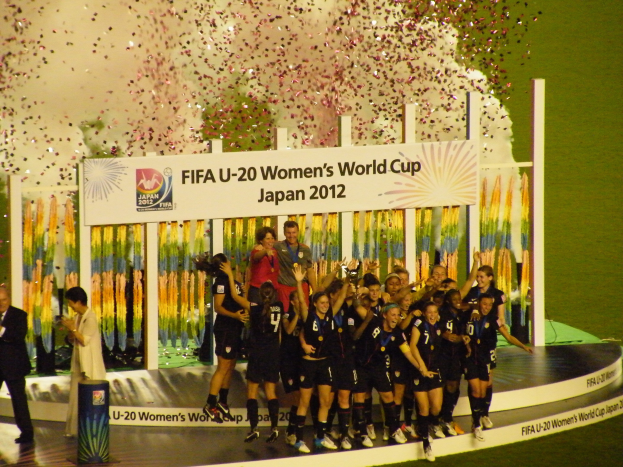 Gruppe von Frauenfußballspielerinnen in schwarzen Uniformen, die auf einer Bühne umgeben von einer Menge feiern, mit einem dekorierten Hintergrund und grasbewachsenem Boden.