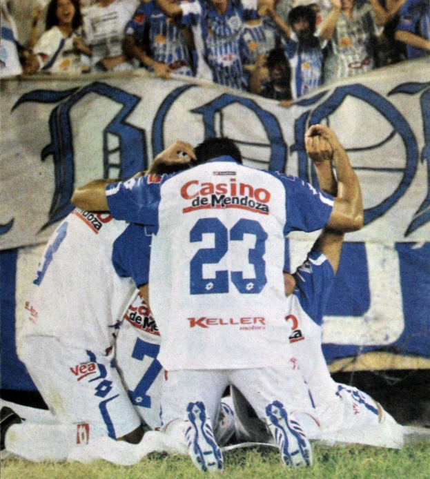 Gruppe von Füßtballspielern, die auf dem Boden vor einer jubelnden Menge mit einem Banner im Hintergrund feiern.