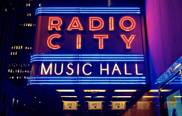 Hell erleuchtetes Radio City Music Hall in New York City mit hohen Gebäuden im Hintergrund, das eine 'Radio City'-Leuchtschrift und zusätzliche Schilder auf der rechten Seite zeigt.