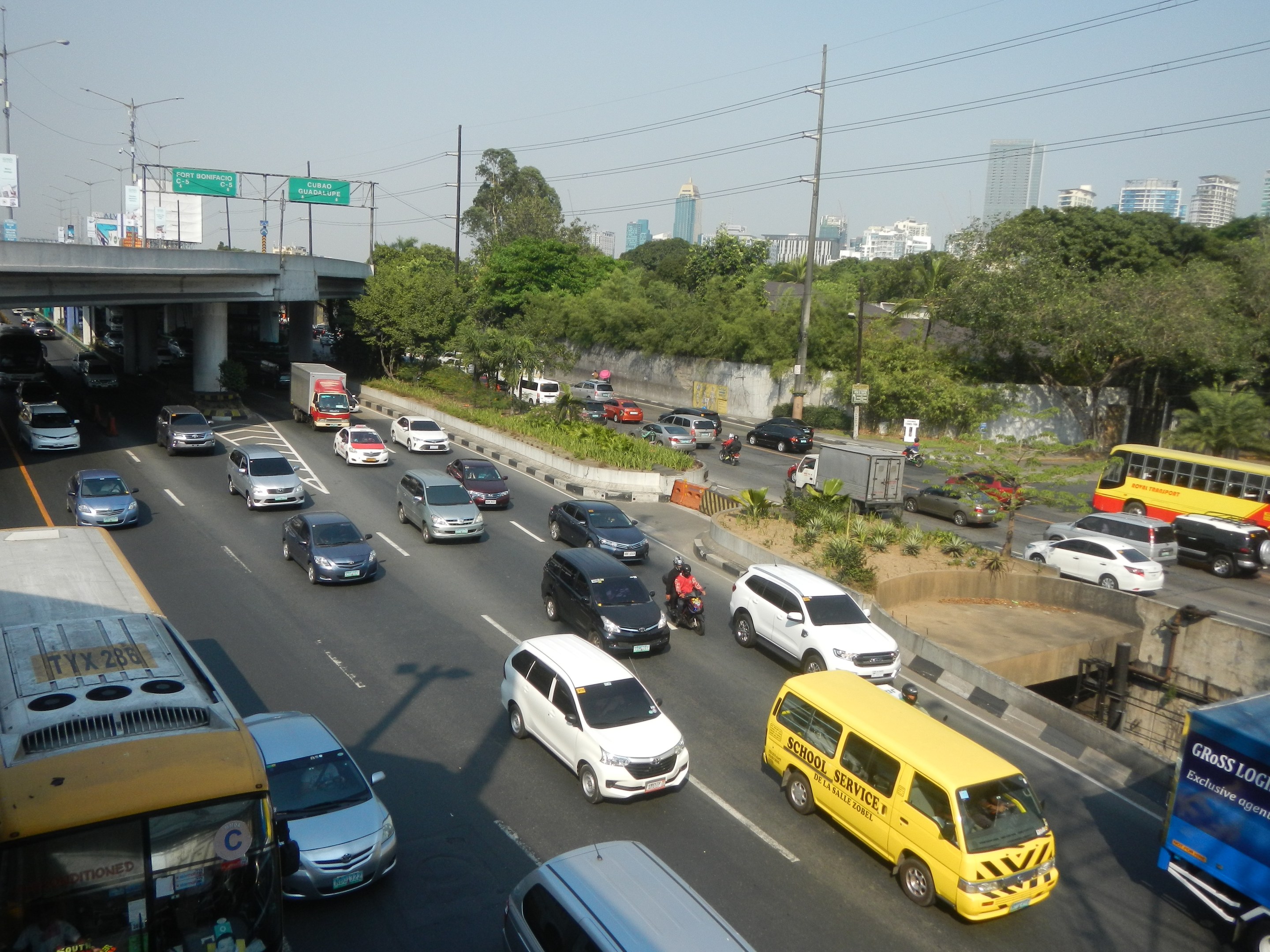 Eine vielbefahrene Autobahn mit Autos, Bussen, Motorrädern, Pflanzen, Bäumen, Strommasten, Schildern, einer Brücke, Laternen und Gebäuden unter einem klaren blauen Himmel.