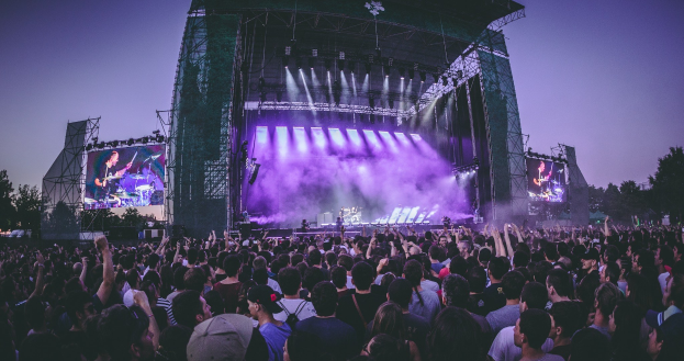 Eine große Menge Menschen bei einem Musikfestival schaut auf einer beleuchteten Bühne mit Instrumenten und Bildschirmen, die vor einem Gebäude, Bäumen und einem klaren Himmel stehen.