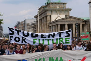 Eine Gruppe von Sch├╝lern marschiert in Berlin, mit einer bunt bemalten Fahne, auf der "Students for Future" steht, im Hintergrund sind Geb├Ąude, B├Ąume und Himmel zu sehen.