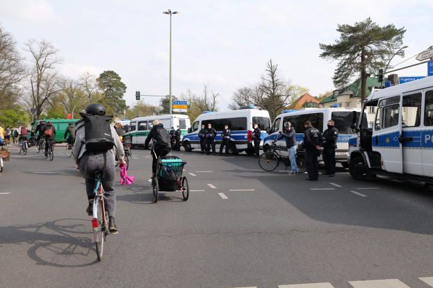 Eine Gruppe von Menschen, die auf Fahrrädern eine Straße entlangfahren, mit Polizeiwagen, anderen Fahrzeugen, Bäumen und Gebäuden im Hintergrund.