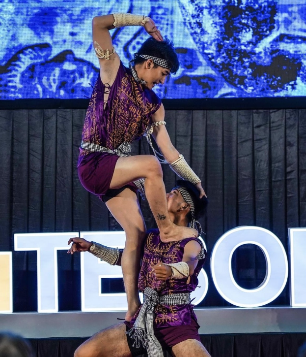 Ein Mann und eine Frau in lila Kleidern führen Akrobatik auf der Bühne auf, wobei der Mann die Frau in seinen Armen hält, während das Publikum von unten zusieht und ein schwarzer Vorhang und ein Bildschirm im Hintergrund zu sehen sind.