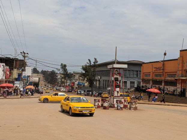 Ein gelbes Taxi fährt auf einer belebten Stadtstraße mit Gebäuden an den Seiten, während Fußgänger mit Regenschirmen auf dem Bürgersteig gehen, unter einem bewölkten Himmel und Stromleitungen.