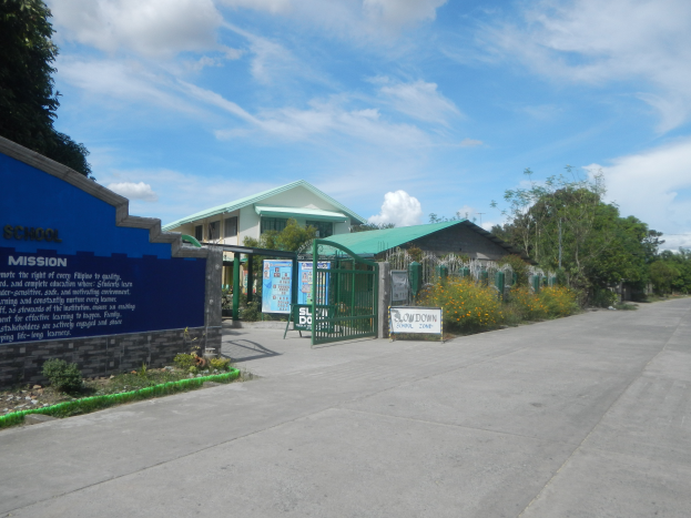 Der Eingang einer Schule mit einem blauen Schild, umgeben von Häusern, Bäumen, Pflanzen, Blumen und einer Straße, unter einem Himmel voller weißer, flauschiger Wolken.