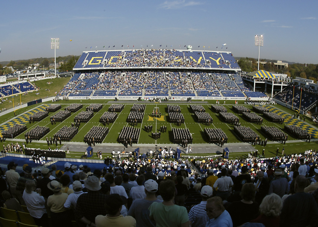 Eine große Menschenmenge schaut einem Fußballspiel in einem Rasenstadion unter einem klaren blauen Himmel zu, mit Laternen, Bäumen und Gebäuden im Hintergrund.