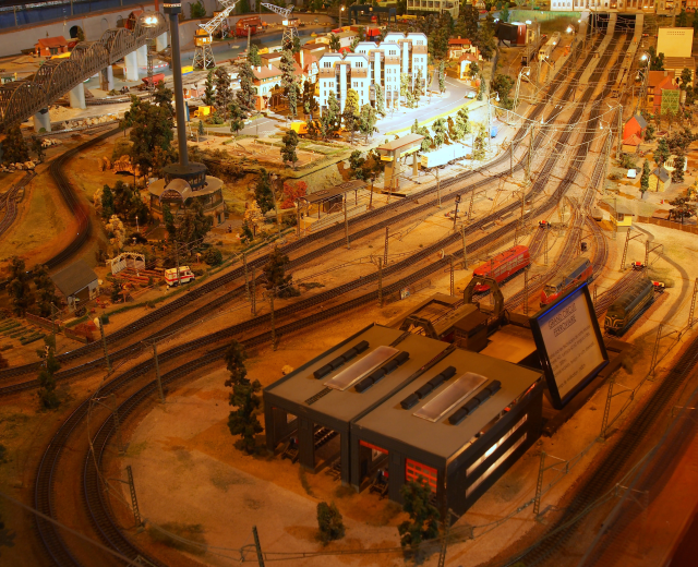 Eine Modellbahnanlage in einem Museum mit Gebäuden, Bäumen, Eisenbahnschienen, Fahrzeugen, Pfählen, Lichtern, Platten und anderen Gegenständen, die eine lebhafte Stadtkulisse schaffen.
