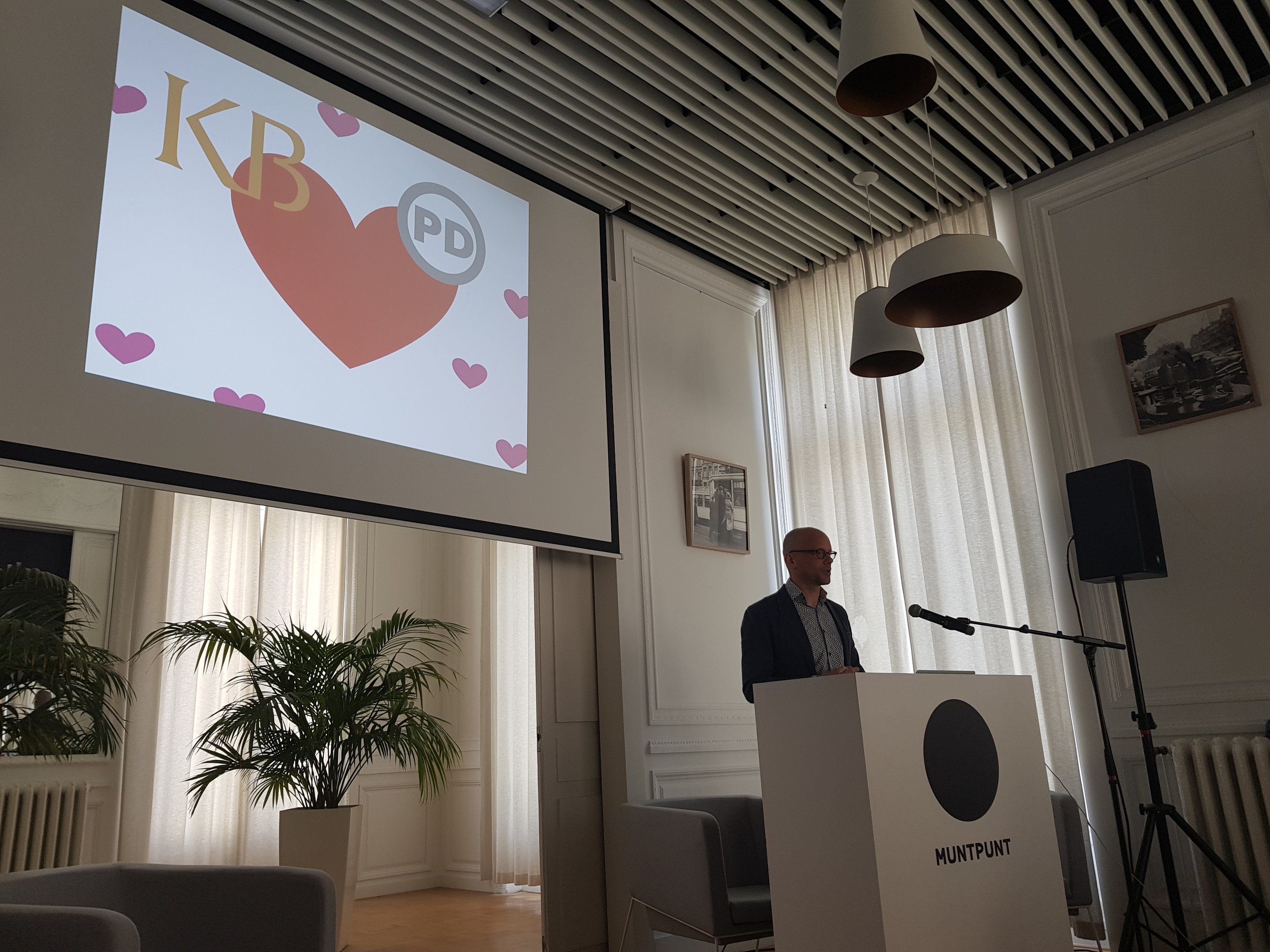 Mann an einem Rednerpult, der über Geschlechtergerechtigkeit pr├Ąsentiert, mit Projektionsleinwand, Mikrofon, Lautsprecher, Stühlen, Topfpflanze, gerahmten Fotos an der Wand, Vorh├Ąngen und Deckenleuchten.