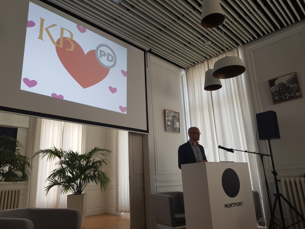 Mann an einem Rednerpult, der über Geschlechtergerechtigkeit pr├Ąsentiert, mit Projektionsleinwand, Mikrofon, Lautsprecher, Stühlen, Topfpflanze, gerahmten Fotos an der Wand, Vorh├Ąngen und Deckenleuchten.