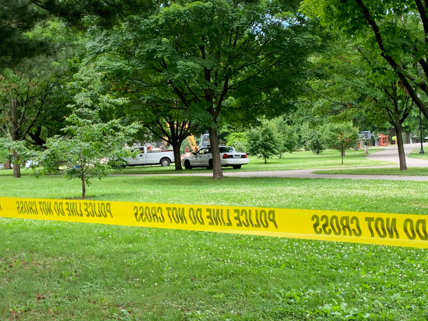 Ein grasbewachsener Bereich mit gelbem Polizeiabsperrband in der Mitte, umgeben von Bäumen und Fahrzeugen auf der Straße, wahrscheinlich die Nachwirkung eines Schusses in einem Park.