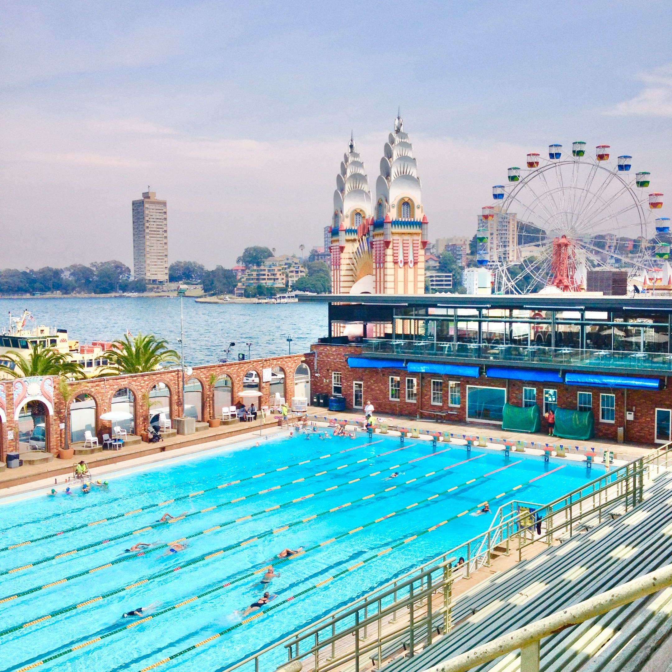 Großes Freibad mit Menschen, umgeben von Geländern und Bänken, mit einem Riesenrad, Gebäuden, Bäumen und bewölktem Himmel im Hintergrund.