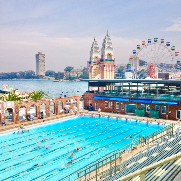 Großes Freibad mit Menschen, umgeben von Geländern und Bänken, mit einem Riesenrad, Gebäuden, Bäumen und bewölktem Himmel im Hintergrund.