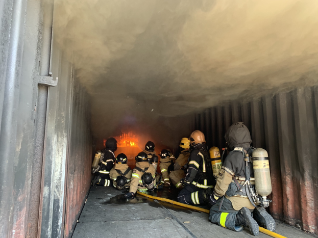 Feuerwehrleute mit Helmen und Sauerstoffflaschen arbeiten in einem raucherfüllten Tunnel, mit einem sichtbaren Feuer im Hintergrund und einer Wand auf der linken Seite.