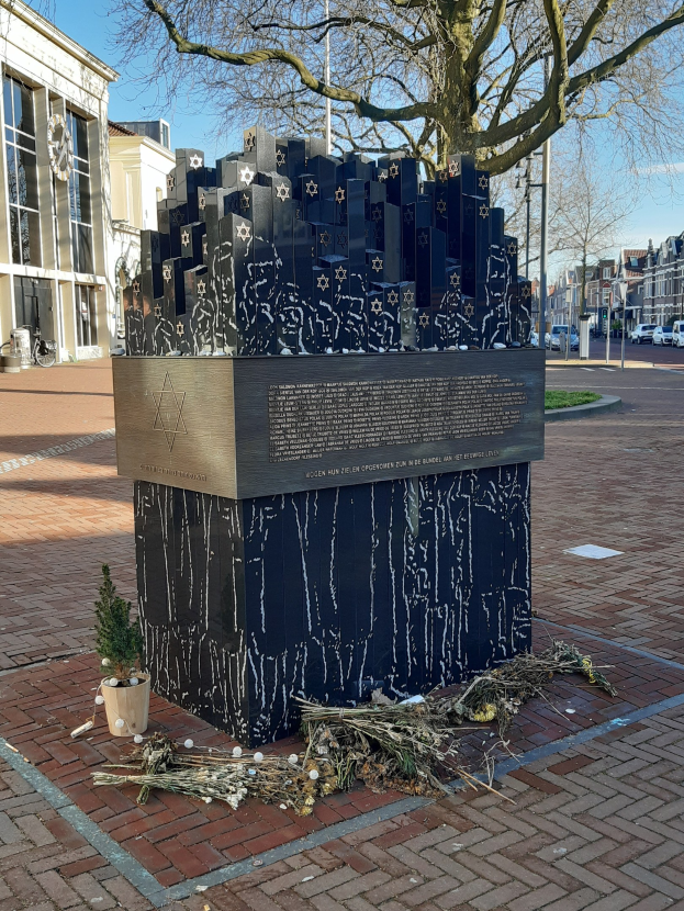 Denkmal in einem Stadtplatz für Holocaust-Opfer, mit einem Baum davor, einem Topfpflanze auf dem Boden, verstreuten trockenen Blättern und Gebäuden, Fahrzeugen und Bäumen im Hintergrund.