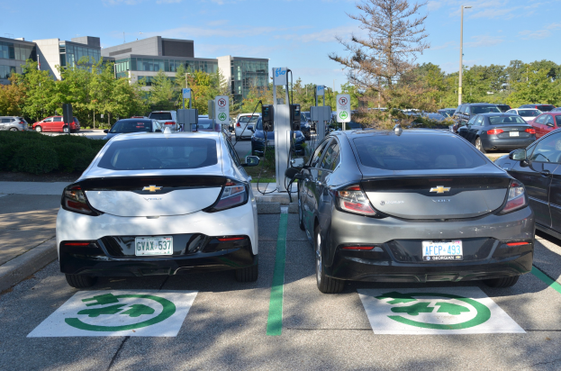 Zwei Elektroautos nebeneinander auf einem Parkplatz mit Bäumen, Pflanzen, Pfählen und Gebäuden im Hintergrund bei bewölktem Himmel.