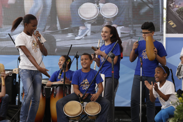 Kinder, die auf der Bühne mit Musikinstrumenten spielen, eine Frau mit Mikrofon und ein Banner im Hintergrund.