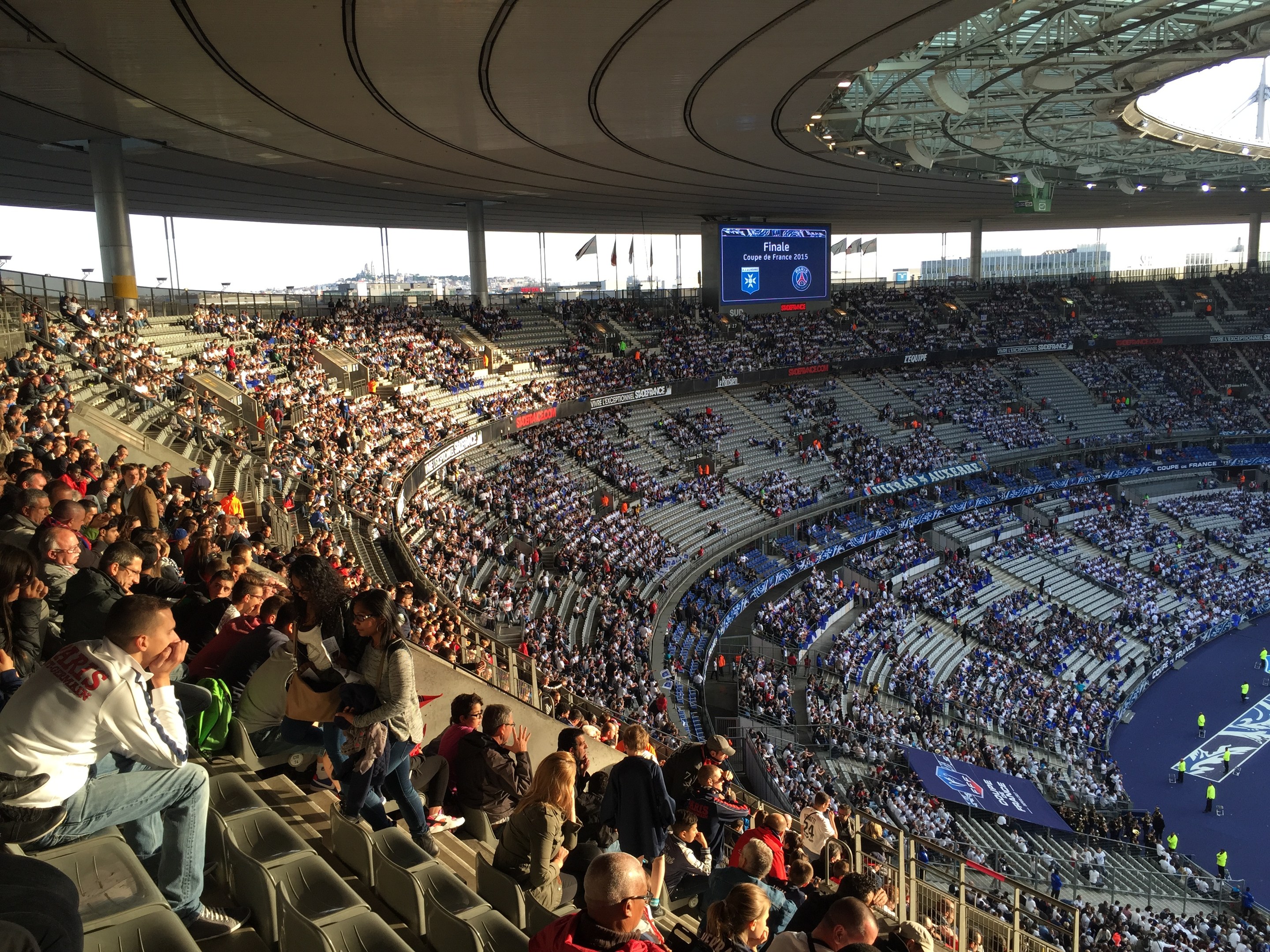 Eine große Menschenmenge in einem Stadion schaut ein Fötballspiel zu, mit einer Bühne auf der rechten Seite, Fahnen, Masten, einem Bildschirm und dem Allianz Stadion in München im Hintergrund.