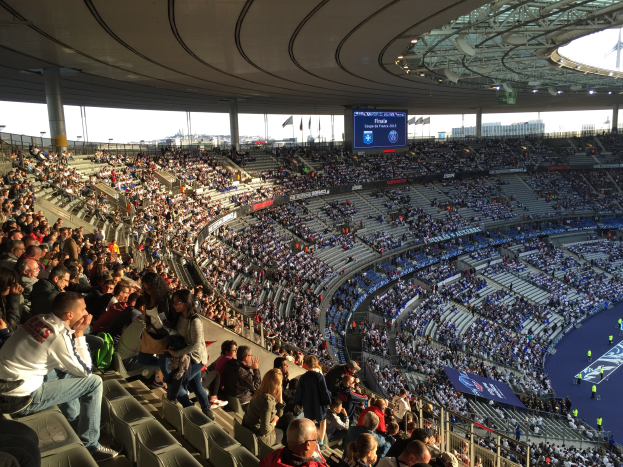 Eine große Menschenmenge in einem Stadion schaut ein Fötballspiel zu, mit einer Bühne auf der rechten Seite, Fahnen, Masten, einem Bildschirm und dem Allianz Stadion in München im Hintergrund.