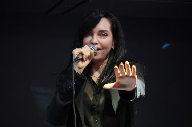 Eine Frau in einem schwarzen Kleid singt in ein Mikrofon auf einer Bühne, ihre Augen geschlossen und ihr Gesichtsausdruck zeigt Emotion und Fokus, beleuchtet von einem hellen Licht.