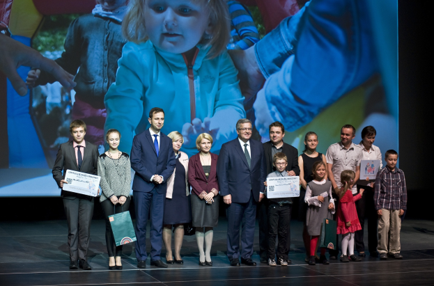 Eine Gruppe von Menschen auf einer Bühne mit Taschen und Urkunden, mit einem Bildschirm, der ein Bild eines Kindes und andere im Hintergrund anzeigt.