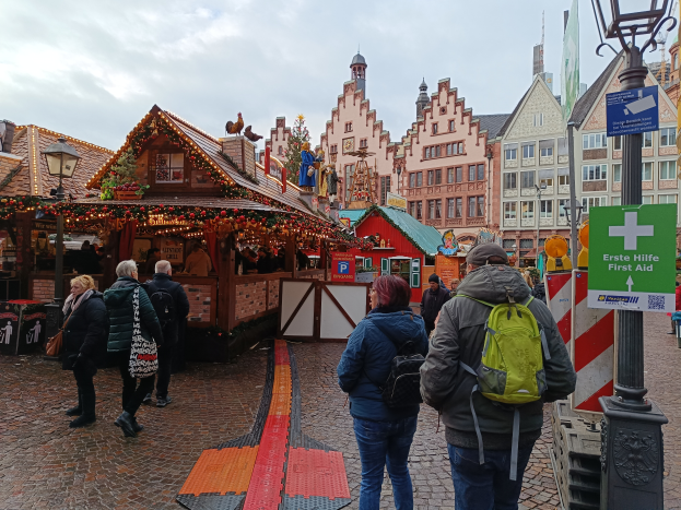Eine Gruppe von Menschen geht eine Kopfsteinpflasterstraße neben einem Weihnachtsmarkt in Nürnberg, Deutschland, entlang mit Laternenmasten, Texttafeln und Gebäuden mit Fenstern im Hintergrund bei einem bewölkten Himmel.