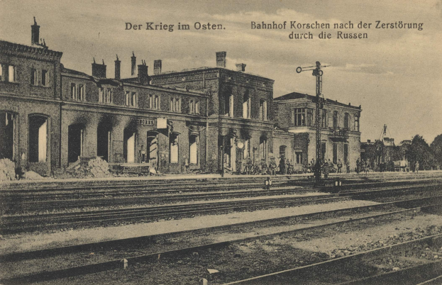 Schwarzes und weiße Foto der Bahnstation Korschen in Ostern, Deutschland, mit Eisenbahnschienen, Stationsgebäuden, Menschen, Bäumen und einem Himmel-Hintergrund mit dem Text "Bahnhof Korschen in Ostern, Deutschland" oben und unten.