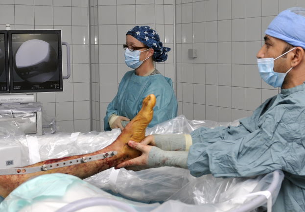 Zwei Chirurgen in Operationskleidung und Masken operieren am Bein eines Patienten in einem Operationssaal, mit medizinischer Ausrüstung und einer Wand im Hintergrund.