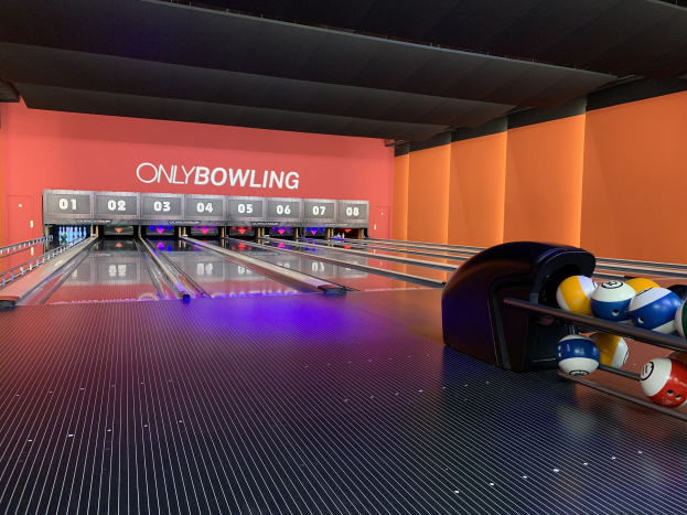 Eine Bowlinganlage mit Bowlingkugeln rechts, Pins in der Mitte und einer Wand mit Text im Hintergrund, unter einer sichtbaren Decke.