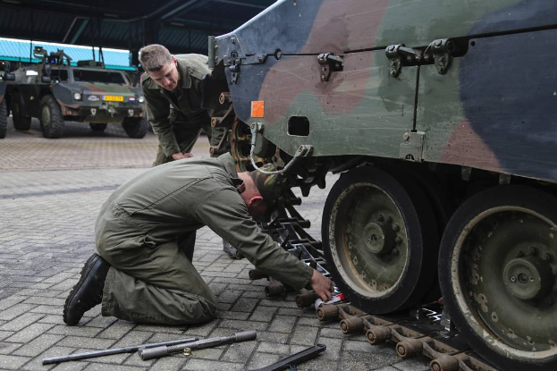 Zwei Männer in militärischen Uniformen arbeiten an einem Militärfahrzeug in einer Garage, umgeben von Werkzeugen, mit mehreren anderen Militärfahrzeugen und einer Hütte im Hintergrund.
