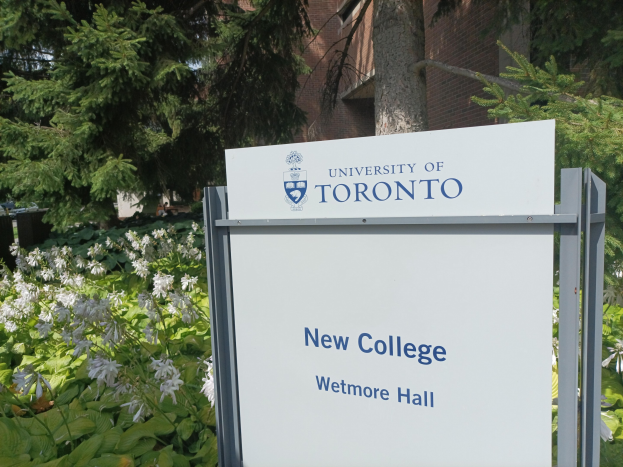 Das Bild zeigt das neue College der Universität Toronto am Wetmore Hall, mit einem Schild vor dem Gebäude, umgeben von grünem Rasen, bunten Blumen, hohen Bäumen und einem hellblauen Himmel.