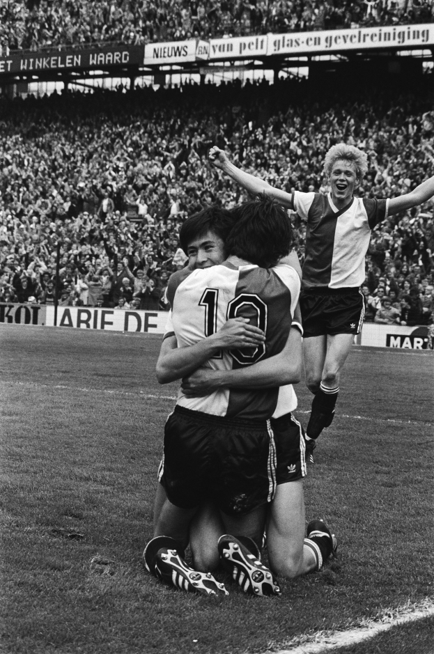 Drei Fußballspieler umarmen sich auf dem Boden nach einem Tor, mit einer jubelnden Menge und schwenkenden Fahnen im Hintergrund, auf einem Schwarz-Weiß-Bild.