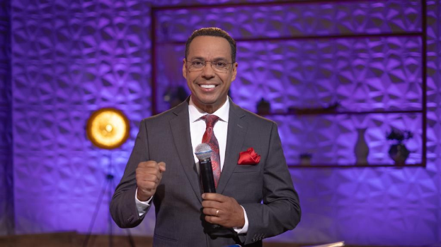 Stephen Colbert, Moderator von *The Daily Show with Trevor Noah*, steht auf einer Bühne und hält ein Mikrofon in der Hand, trägt einen Anzug, eine Krawatte und eine Brille, mit einem Scheinwerfer und Ständer hinter ihm.