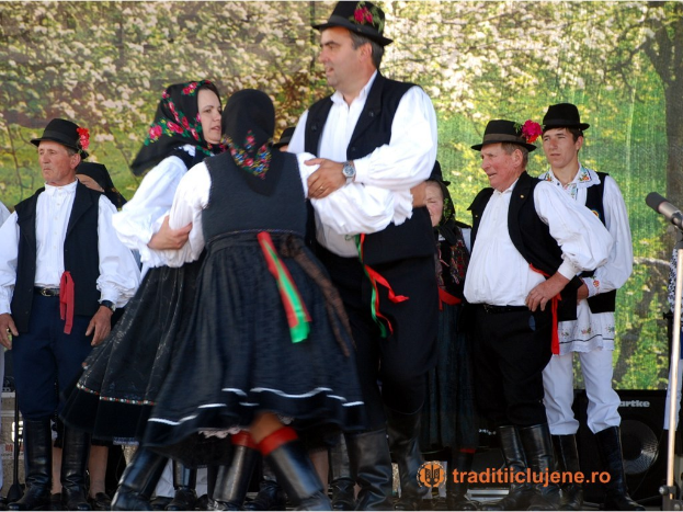 Gruppe von Menschen in traditioneller ungarischer Kleidung tanzt auf einer Bühne mit Mikrofonen, Lautsprechern und Bäumen im Hintergrund; Text unten.