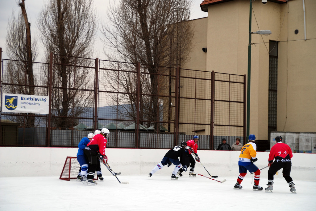 Menschen beim Eishockeyspielen auf einer Eisfläche mit Gebäuden, Bäumen, einer Straßenlaterne, einem Namensschild und Zäunen im Hintergrund bei klarem Himmel.