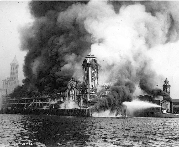 Schwarz-weiß-Foto eines großen Gebäudes, das in Flammen steht und von dem Rauch aufsteigt, mit einem Gewässer im Vordergrund und anderen Gebäuden und dem Himmel im Hintergrund.