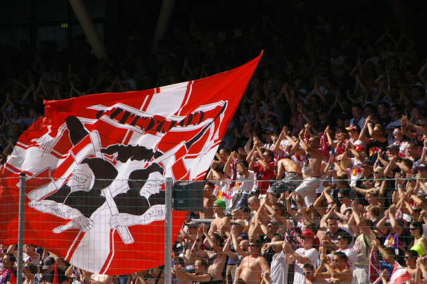 Große Menschenmenge in einem Stadion mit einer rot-weißen Fahne, Maschendrahtzaun im Vordergrund und Säulen im Hintergrund vor dunklem Hintergrund