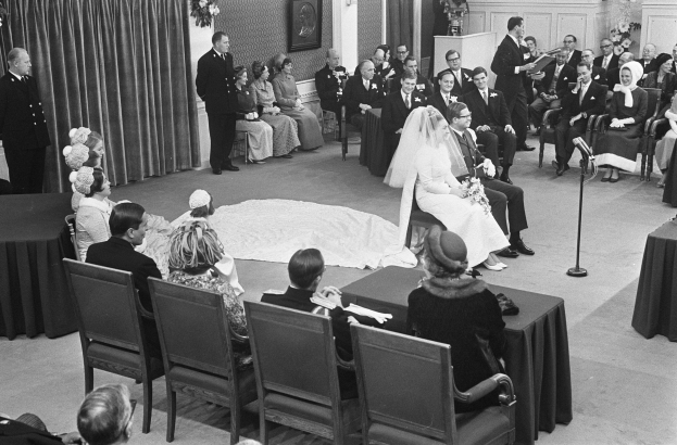 Schwarze und weiße Fotografie einer Hochzeitszeremonie mit Menschen, die sitzen und stehen, einem Mann mit Mikrofon, einer Frau mit Blumenstrauß, einem Podium mit Mikrofon, Blumenschmuck, einem Vorhang, einem Rahmen an der Wand und einer Tür im Hintergrund.