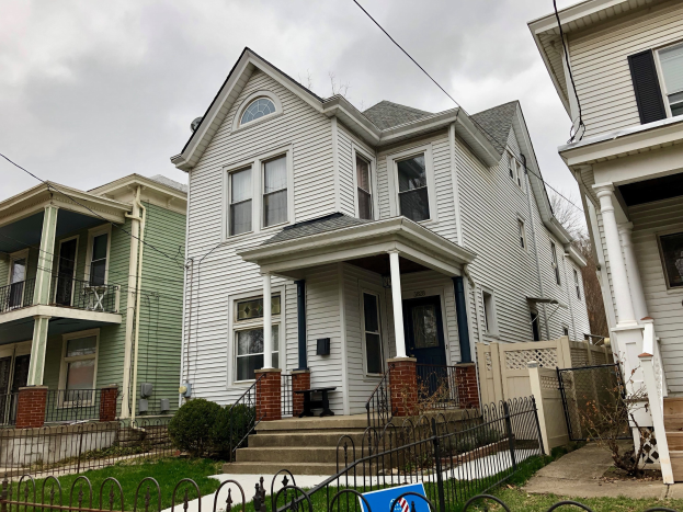 Reihe von Häusern mit Fenstern, Türen, Säulen, Geländern, Treppen und Dächern, umgeben von einem Metallzaun, Gras, Pflanzen, Bäumen und einem bewölkten Himmel, mit einem Mietschild am Boden, das die Adresse und die Details der Liste anzeigt.