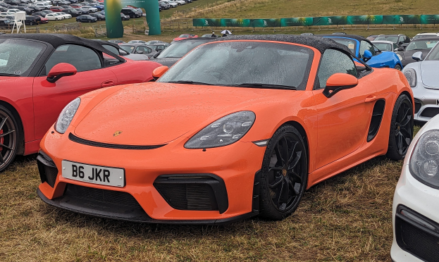 Ein silberner Porsche 718 Boxster GTS auf dem Goodwood Festival of Speed zusammen mit anderen Autos, einem Banner, Bäumen und einem klaren blauen Himmel im Hintergrund.