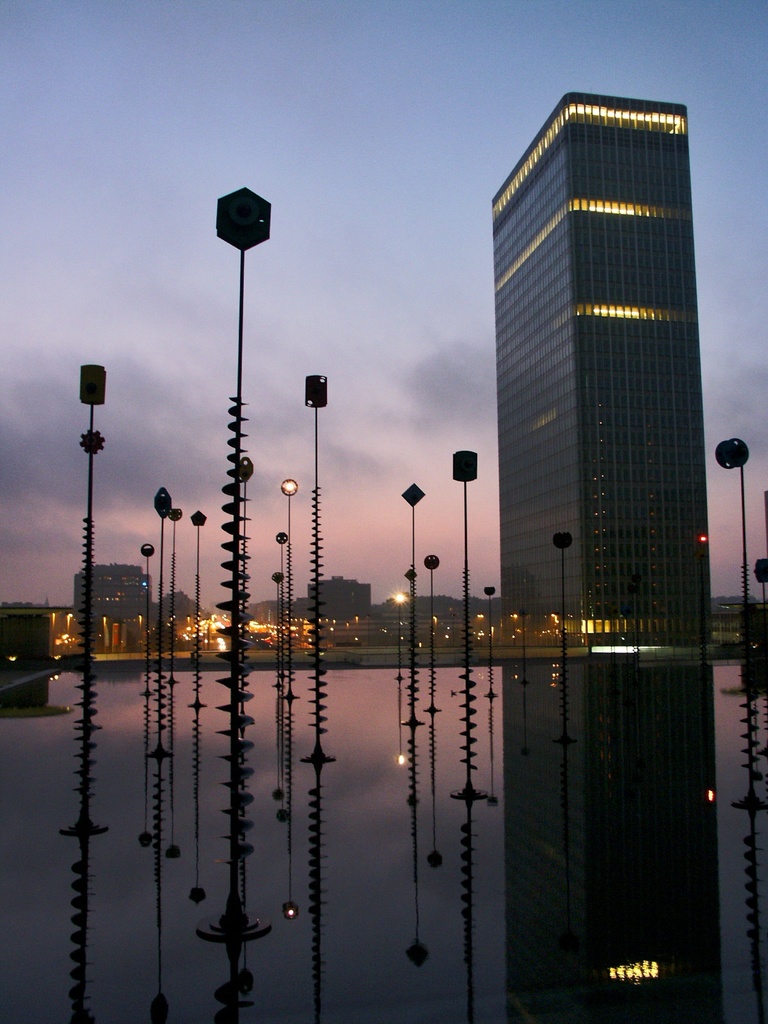 Mehrere Masten und Gebäude mit Lichtern unter einem bewölkten Himmel, mit ihren Spiegelungen
