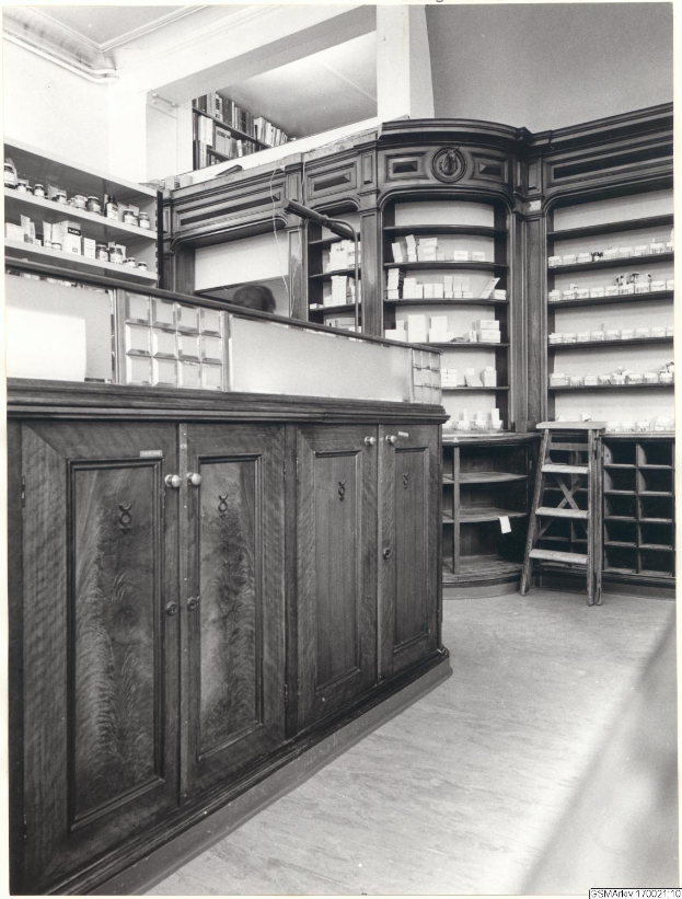 Ein Schwarz-Weiß-Foto eines Apothekeninneren mit einer Theke, Schränken, einer Leiter und Regalen voller verschiedener Gegenstände, mit Text oben.