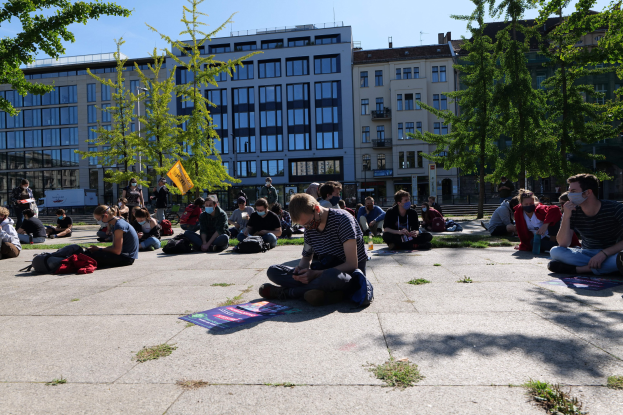 Eine Gruppe von Menschen sitzt vor einem Gebäude auf dem Boden während einer Demonstration in Berlin, einige tragen Masken mit Taschen und Gegenständen, die herumliegen, unter einem klaren blauen Himmel mit Bäumen und Gebäuden im Hintergrund.